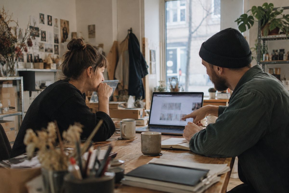 Zwei Personen sitzen an einem großen Holztisch in einem Atelier und besprechen gemeinsam eine Website auf dem Laptop